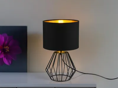 A black table lamp with a gold interior is placed on a white surface next to a framed picture of a purple flower.
