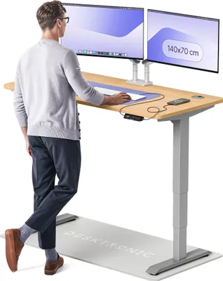 A man is standing at a height-adjustable desk with two monitors, using a mouse and keyboard. There is an anti-fatigue mat on the floor.