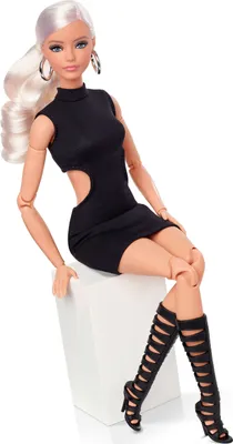 A Barbie doll is sitting on a white cube, wearing a black dress and black gladiator boots.