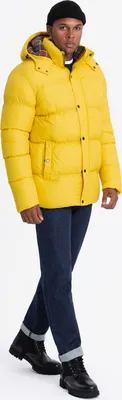 A man wearing a yellow puffer jacket and blue jeans, standing against a white background.