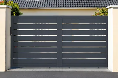 A modern, dark gray aluminum sliding gate with horizontal slats, set between two light-colored pillars in front of a house with a tiled roof.