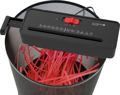 A black paper shredder is placed on top of a black trash bin, shredding red paper into strips.