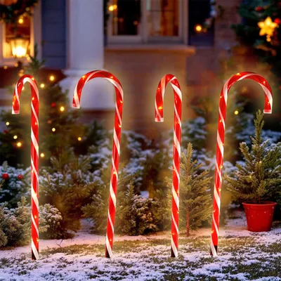 Four illuminated candy canes are placed in the snow in front of a house with Christmas lights.