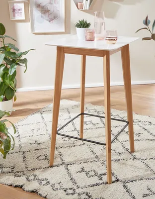 A modern bar table with wooden legs and a white top, placed on a patterned rug in a well-lit room.