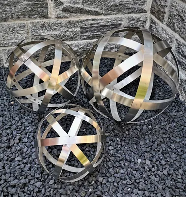 Three metallic decorative spheres are placed on a bed of dark gravel in front of a stone wall.