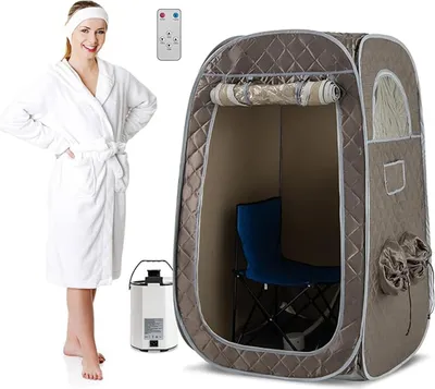 A woman in a white robe stands next to a portable steam sauna tent, a steam generator, and a remote control.