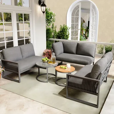 A patio set with gray cushions and dark frames is arranged on a light-colored rug on a tiled patio, with a white arched doorway and greenery in the background.