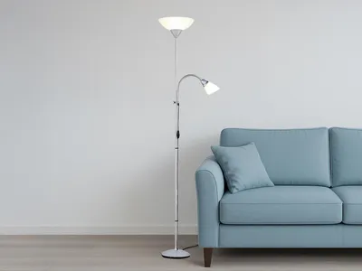 A silver floor lamp with two lights stands next to a light blue sofa against a white wall.