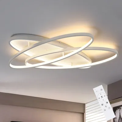 A modern white LED ceiling light with multiple interlocking rings, illuminated and mounted on a white ceiling. A remote control is shown in the bottom right corner.