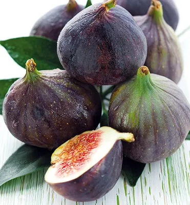 A close-up of several dark purple figs, with one fig cut in half to reveal its red interior, arranged on a white surface with green leaves.