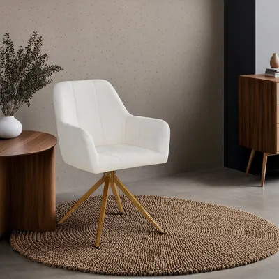 A white armchair with gold legs sits on a round brown rug next to a wooden side table with a plant in a white vase. A wooden cabinet is visible in the background.
