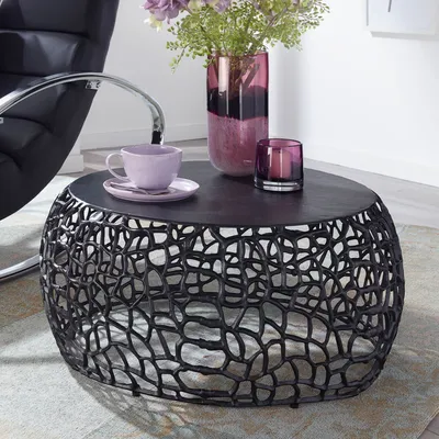A round coffee table with a decorative base, set in a living room with a chair and decorative items on top.