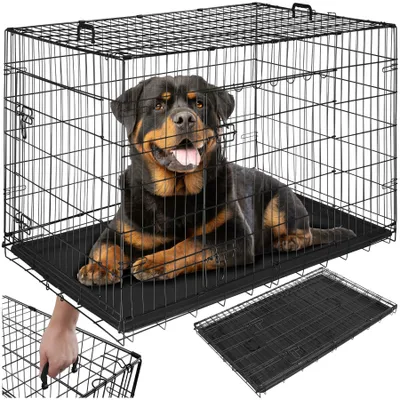 A Rottweiler dog is lying inside a black wire dog crate. The crate is shown from the front and slightly to the side. A second, disassembled crate and a tray are shown in the foreground.