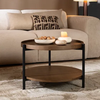 A round, two-tiered coffee table with a dark oak finish and black metal legs, placed on a rug in a living room setting.