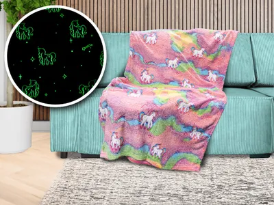 A fluffy blanket with a unicorn and rainbow pattern is draped over a teal sofa. A circular inset shows a close-up of the blanket's glow-in-the-dark unicorn pattern on a black background.