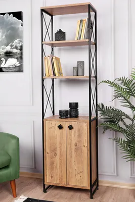 A tall, narrow bookshelf with a wooden cabinet at the bottom, set against a white wall with a green chair and a plant visible.