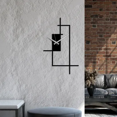 A modern black metal wall clock with a minimalist design is mounted on a textured white wall, with a brick wall and a sofa visible in the background.
