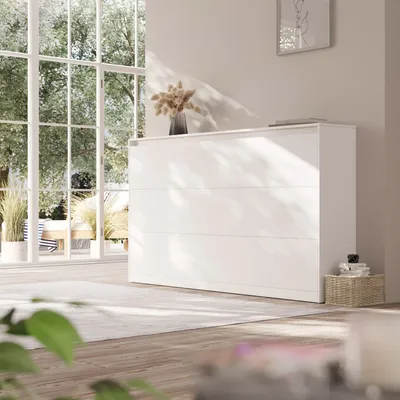 A white dresser with three drawers is placed in a bright room with a large window and outdoor scenery visible.
