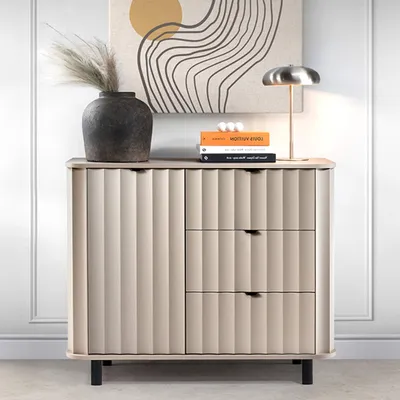 A modern beige sideboard with fluted fronts and black legs, styled with a vase of dried grass, books, and a lamp on top, against a white wall with molding.