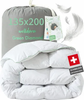 A folded white duvet with green trim, presented in a grey drawstring bag with product information printed on it. Several white feathers are scattered around the image.