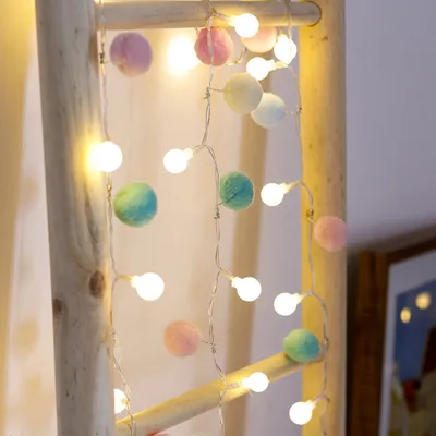 A string of fairy lights with pom-poms and bulbs draped over a wooden ladder.
