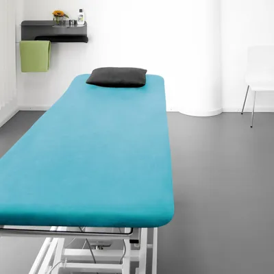 A treatment table covered with a turquoise sheet, with a black pillow and a green towel hanging on a shelf.