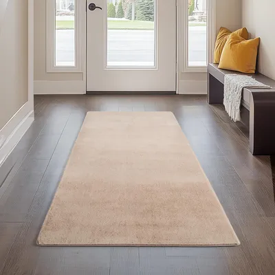 A beige rug is laid out on a dark wood floor in a hallway, with a white door and a dark bench with yellow pillows in the background.
