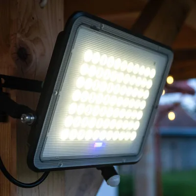 A solar floodlight with a motion sensor mounted on a wooden post, with a blurred background of houses and lights.