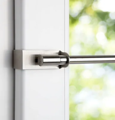 A close-up view of a tension rod attached to a white window frame, with blurred green foliage visible through the window.