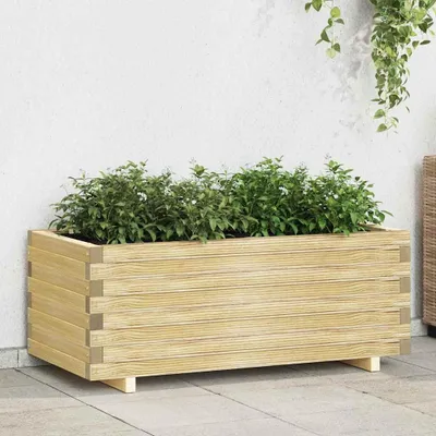 A rectangular wooden planter box filled with green plants, placed on a patio against a white wall.