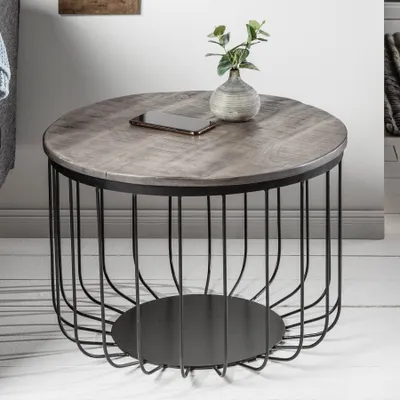 A round coffee table with a wooden top and a black metal cage-like base, placed on a light-colored floor.