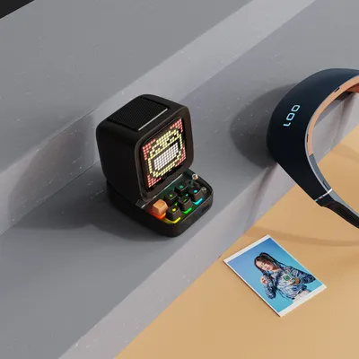 A small retro-style speaker with a pixelated screen and colorful keyboard keys sits on a gray surface next to a pair of headphones and a small photo of a person.