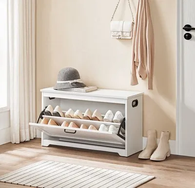 A white shoe cabinet with a hat and a book on top, placed in a hallway next to a door and a window with curtains. A beige cardigan and a white handbag are hanging on the wall.