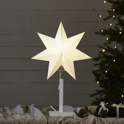 A lit paper star decoration on a stand, placed on a table with Christmas gifts and a decorated Christmas tree in the background.