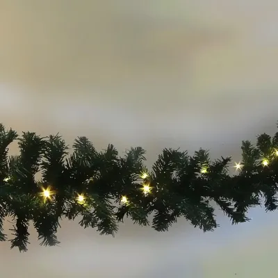 A close-up shot of a green Christmas garland with small, warm white lights, draped against a light gray background.