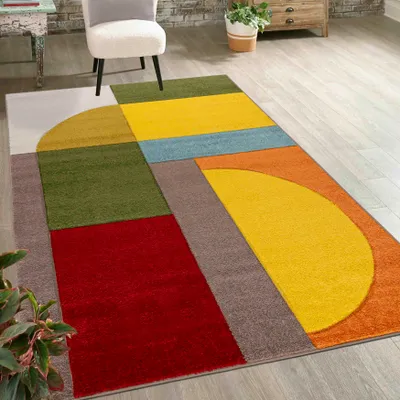 A colorful geometric rug is laid out on a wooden floor in a room setting, with a chair and a plant visible.
