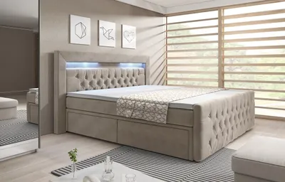 A bedroom scene featuring a large, upholstered bed with a tufted headboard and integrated LED lighting. The bed has storage drawers and is dressed with bedding and a throw. A large window with blinds is visible in the background, and a mirror reflects part