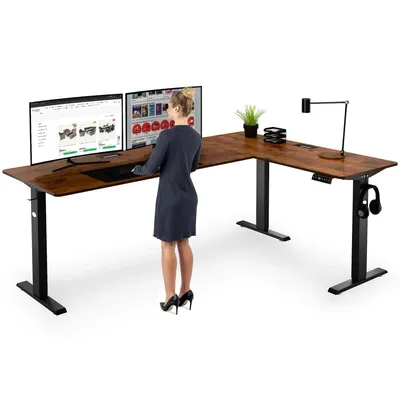 A woman stands at a large L-shaped standing desk with two monitors, a plant, and a lamp.