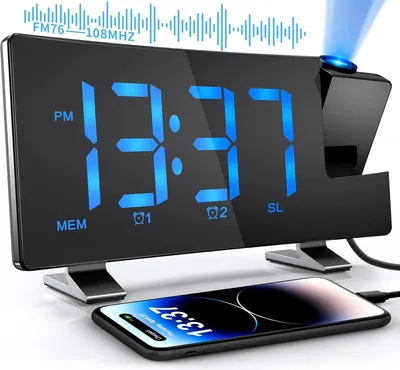 A black digital alarm clock with a blue LED display and a blue projection beam is shown on a white surface, with a smartphone charging in front of it.
