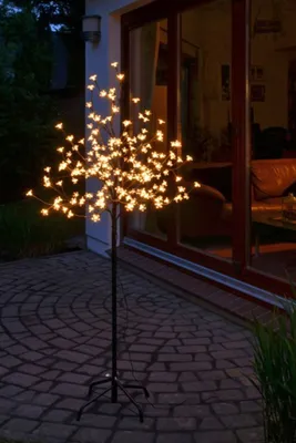 A decorative LED cherry blossom tree is lit up at night on a paved patio next to a house with large glass doors.