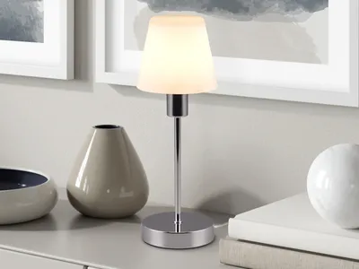A table lamp with a glass shade sits on a dresser next to decorative vases and books, with artwork on the wall behind.