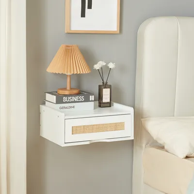 A white floating shelf with a drawer is mounted on a gray wall next to a bed. The shelf holds a lamp, books, and a vase with flowers.
