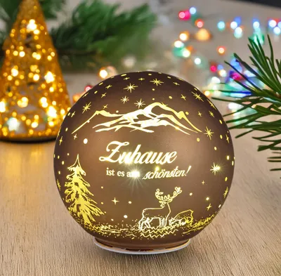 A decorative glass sphere with LED lights, featuring a winter mountain scene and text, placed on a wooden surface with Christmas lights and pine branches in the background.