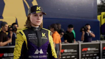 A female racing driver in a yellow and purple racing suit and cap stands on a podium with other people and cameras in the background.