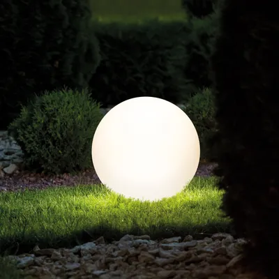 A glowing spherical solar light sits on a grassy lawn at night, surrounded by bushes and gravel.