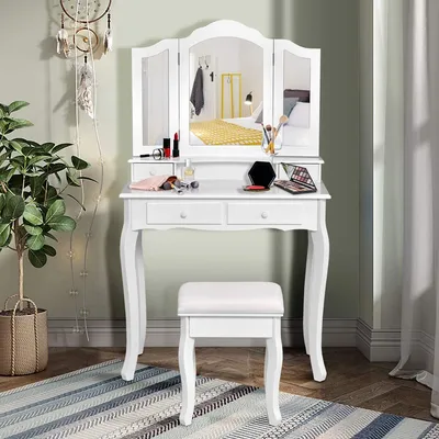 A white vanity table with a tri-fold mirror, stool, and makeup accessories, set in a room with a plant and a rug.