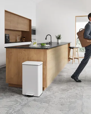 A person is walking out of a modern kitchen with a briefcase, while a white trash can is placed in the foreground.