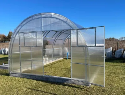 A greenhouse with its doors open, set up on a grassy lawn under a clear blue sky.