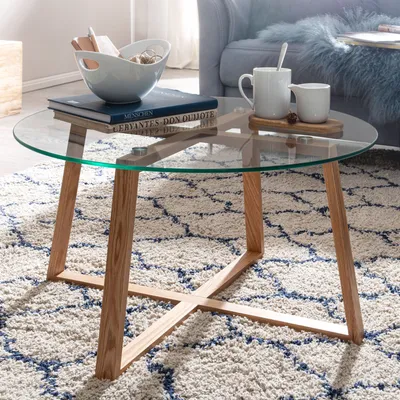 A round glass coffee table with wooden legs, decorated with books, a bowl, and mugs, placed on a patterned rug in a living room setting.