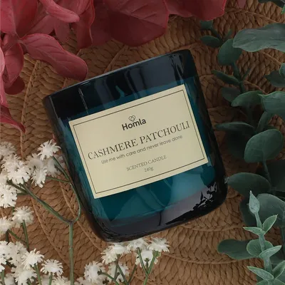 A scented candle with a label is placed on a woven mat, surrounded by flowers and greenery.